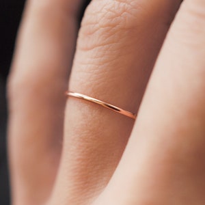 K&ouml;nnte beinhalten: Ein zarter, d&uuml;nner Ring aus Ros&eacute;gold wird an einem Finger getragen. Der Ring ist ein schlichtes Band, das ein minimalistisches Design zeigt. Der Fokus liegt auf dem Schmuck, der seine elegante Schlichtheit und den warmen Farbton des Metalls hervorhebt.