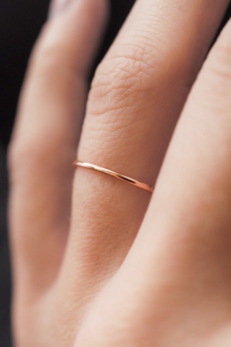 K&ouml;nnte beinhalten: Ein zarter, d&uuml;nner Ring aus Ros&eacute;gold wird an einem Finger getragen. Der Ring ist ein schlichtes Band, das ein minimalistisches Design zeigt. Der Fokus liegt auf dem Schmuck, der seine elegante Schlichtheit und den warmen Farbton des Metalls hervorhebt.