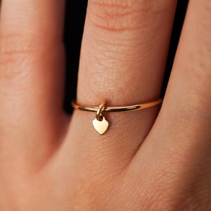May include: A gold ring with a small heart charm dangling from it. The ring is worn on a finger.