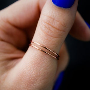 K&ouml;nnte beinhalten: Nahaufnahme von zwei d&uuml;nnen Ringen aus Ros&eacute;gold. Die Ringe sind auf einem Finger gestapelt, mit leuchtend blauem Nagellack im Hintergrund. Die Ringe sind zart und minimalistisch.