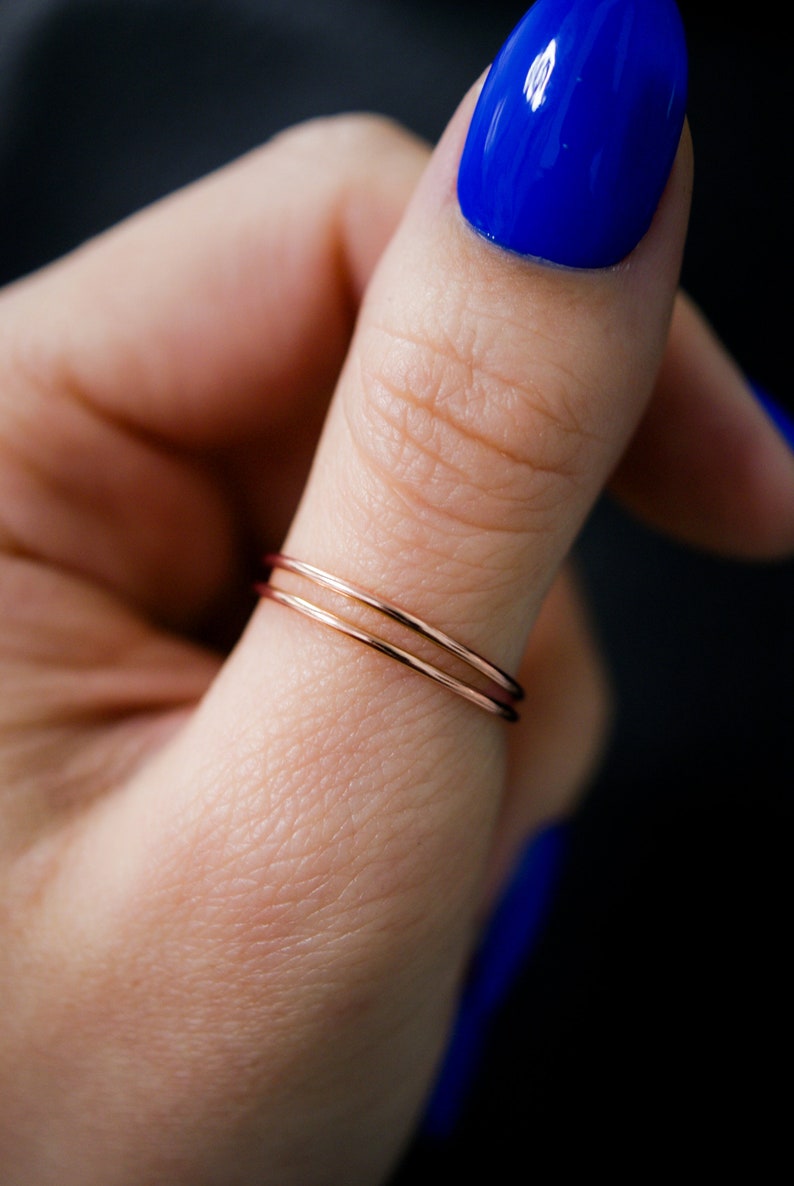 K&ouml;nnte beinhalten: Nahaufnahme von zwei d&uuml;nnen Ringen aus Ros&eacute;gold. Die Ringe sind auf einem Finger gestapelt, mit leuchtend blauem Nagellack im Hintergrund. Die Ringe sind zart und minimalistisch.