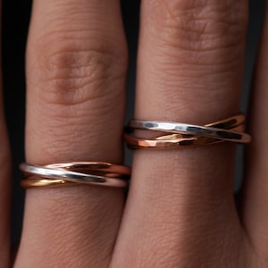 Puede incluir: Primer plano de dos juegos de anillos de tres bandas en oro, plata y oro rosa. Los anillos están entrelazados, creando un diseño único y moderno. Las bandas tienen una textura martillada. Los anillos se muestran en dedos sobre un fondo oscuro.