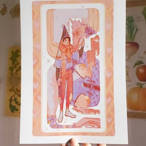 Op de afbeelding: Een ingelijste kunstdruk met een grillige illustratie. Het kunstwerk toont twee figuren, een met een hoed en sjaal, die interageren met een fantastisch wezen. Het kleurenpalet omvat zachte tinten oranje, blauw en roze, met een rand van hartjes.