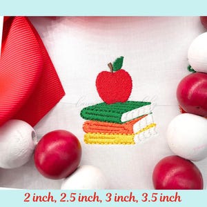 May include: An embroidered design featuring a red apple atop a stack of three books in green, orange, and yellow. The image includes a red bow and decorative red and white spheres. The text at the bottom reads: 2 inch, 2.5 inch, 3 inch, 3.5 inch.