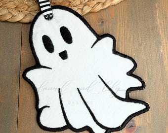 May include: A white ghost-shaped tag with black embroidered outlines and facial features. The tag features a black and white striped ribbon loop at the top. Text on the image reads Ghost Tag In The Hoop and 4 inch, 5 inch, 6 inch.