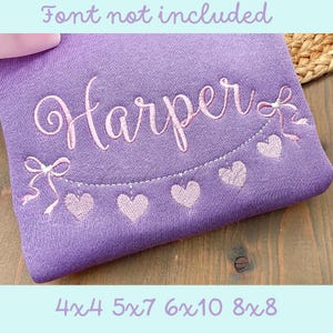 Puede incluir: Sudadera morada con el nombre "Harper" bordado en letras cursivas rosas. Una cinta rosa y una hilera de corazones rosas están bordados debajo del nombre.
