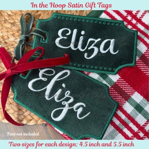 May include: Two dark green satin gift tags embroidered with the name "Eliza" in white script. Each tag has a decorative border and is tied with a velvet ribbon. The tags are 11.4 and 14 cm in size. The text "In the Hoop Satin Gift Tags" is at the top.