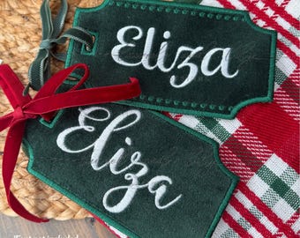 Pode incluir: Duas etiquetas de presente de cetim verde escuro com o nome "Eliza" bordado em branco. Cada etiqueta tem uma borda decorativa e é amarrada com uma fita de veludo. As etiquetas têm 11,4 e 14 cm. O texto "In the Hoop Satin Gift Tags" está no topo.