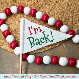 May include: A white pennant flag with the text "I'm Back!" in red and green, on a wooden stick. The flag is surrounded by a garland of red and white wooden beads. The background is a woven straw mat.