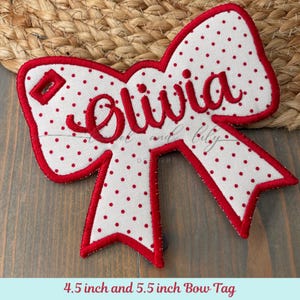 Puede incluir: Una etiqueta blanca con forma de lazo con lunares rojos y borde rojo. El nombre "Olivia" está bordado en rojo en el centro. La etiqueta mide aproximadamente 11,4 y 14 cm.