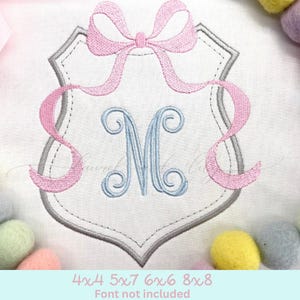 May include: A gray shield with a pink bow and a blue monogram letter M. The shield is outlined in gray and has a scalloped edge. The bow is tied at the top of the shield and the ribbon tails extend down the sides. The monogram is centered in the shield. The image includes the text "4x4 5x7 6x6 8x8 Font not included".