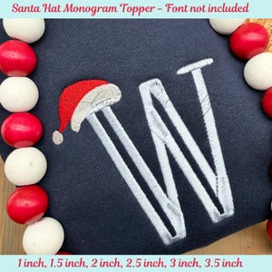 May include: A navy blue monogram topper featuring a white embroidered letter "W" with a Santa hat design. The text "Santa Hat Monogram Topper - Font not included" is at the top. Sizes available: 1 inch to 3.5 inches.