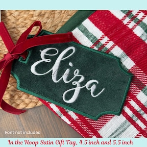May include: A dark green satin gift tag with the name "Eliza" embroidered in white script. The tag is trimmed with green stitching and a red velvet ribbon. The background features a red and white plaid pattern.