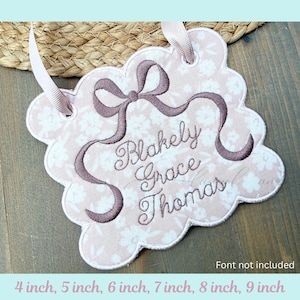 May include: A scalloped fabric plaque featuring a light pink floral pattern with an embroidered dark mauve bow and the name Blakely Grace Thomas in cursive script. The plaque includes two light pink ribbon loops for hanging purposes.
