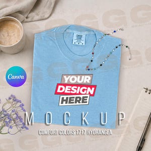 Puede incluir: Maqueta de camiseta Comfort Colors azul claro con el texto "YOUR DESIGN HERE" en un diseño rojo y blanco. La camiseta está doblada y tiene una etiqueta que dice "COMFORT COLORS". Un collar y una taza de café están al lado de la camiseta.