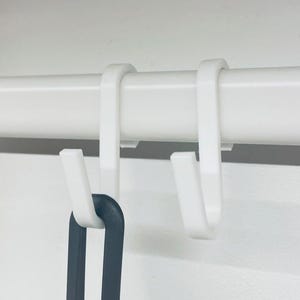 Pode incluir: Dois ganchos de plástico branco pendurados em uma barra branca. Um utensílio preto está pendurado em um dos ganchos.