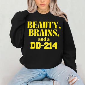 以下が含まれることがあります： 黄色い文字で「BEAUTY, BRAINS, and a DD-214」と書かれた黒いスウェットシャツ。 スウェットシャツはクルーネックで、青いジーンズを履いた人が着ています。