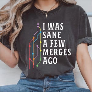 May include: Dark gray t-shirt with the text "I WAS SANE A FEW MERGES AGO" in white. A colorful graphic resembling a circuit board is above the text. The shirt is a casual wear item.