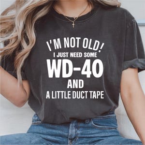 May include: Dark gray t-shirt with the white text "I'M NOT OLD! I JUST NEED SOME WD-40 AND A LITTLE DUCT TAPE." The shirt is a casual style, suitable for everyday wear.
