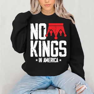 Könnte beinhalten: Schwarzes Sweatshirt mit dem Text "NO KINGS IN AMERICA" in weißer, abgenutzter Schrift. Eine rote Kronengrafik befindet sich über dem Wort "KINGS". Das Sweatshirt wird mit einer blauen Jeans getragen.