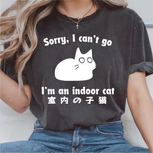 以下が含まれることがあります： 「Sorry, I can't go」の文字と白い猫のイラストがプリントされた濃いグレーのTシャツ。猫の下には「I'm an indoor cat」と日本語のテキスト。カジュアルなスタイルのシャツです。