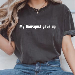 Può includere: T-shirt grigio scuro con la scritta bianca "My therapist gave up." La maglietta è in stile casual, con maniche arrotolate. La modella indossa jeans blu e una collana d'oro.