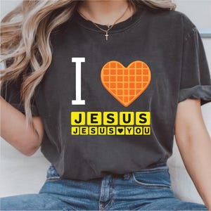 May include: Dark gray t-shirt with the words "I" and a waffle-shaped heart. Below, the text reads "JESUS JESUS YOU" in yellow block letters. The shirt is a casual style.