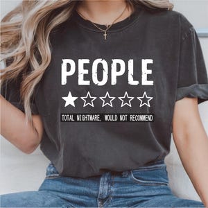 May include: Dark gray t-shirt with the word "PEOPLE" in large white letters above five star shapes. Below the stars, the text reads "TOTAL NIGHTMARE, WOULD NOT RECOMMEND". The shirt is worn by a person.