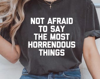 May include: Dark gray t-shirt with the text "NOT AFRAID TO SAY THE MOST HORRENDOUS THINGS" in white block letters. The shirt is a crew neck style and is worn by a person wearing blue jeans.