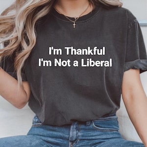 May include: Dark gray t-shirt with the white text "I'm Thankful I'm Not a Liberal." The shirt is a crew neck style and is worn with blue jeans. The image shows the upper body of a person.