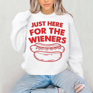 Op de afbeelding: Witte sweatshirt met de rode tekst "JUST HERE FOR THE WIENERS" en een rode hotdog-afbeelding. Een casual kledingstijl.