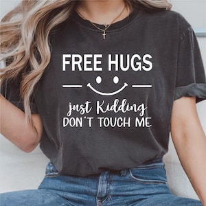 以下が含まれることがあります： 「FREE HUGS」の文字と笑顔のグラフィックがプリントされた濃いグレーのTシャツ。グラフィックの下には「just Kidding DON'T TOUCH ME」と書かれています。Tシャツは人が着ています。