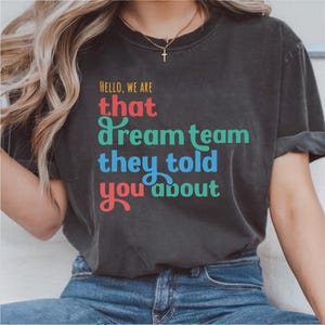Puede incluir: Camiseta gris oscuro con el texto "HELLO, WE ARE that dream team they told you about" en letras coloridas. La camiseta es de cuello redondo y tiene mangas enrolladas.