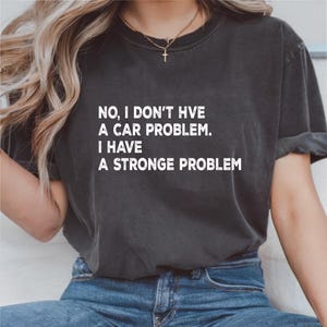 May include: Dark gray t-shirt with the text "NO, I DON'T HVE A CAR PROBLEM. I HAVE A STRONGE PROBLEM" in white. The shirt is worn with blue jeans.