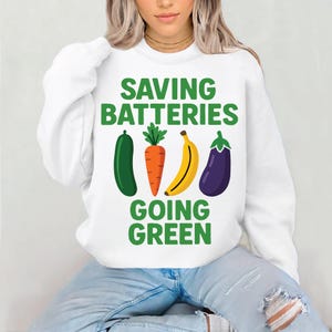 May include: White sweatshirt featuring the text "SAVING BATTERIES GOING GREEN" in green. The design includes illustrations of a green cucumber, orange carrot, yellow banana, and purple aubergine.
