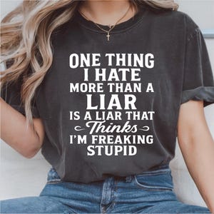 Può includere: T-shirt grigio scuro con la scritta bianca "ONE THING I HATE MORE THAN A LIAR IS A LIAR THAT Thinks I'M FREAKING STUPID". La maglietta è indossata con jeans blu.