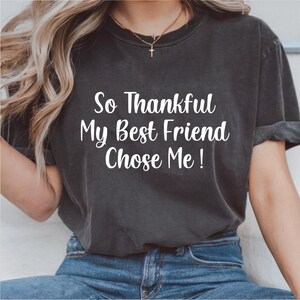May include: Dark grey t-shirt with the text "So Thankful My Best Friend Chose Me!" in white. The shirt is a crew neck style and appears to be made of a soft material. The model is wearing blue jeans.