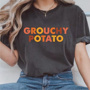 以下が含まれることがあります： オレンジ、赤、黄色の文字で「GROUCHY POTATO」と書かれた濃い灰色のTシャツ。青いジーンズと合わせて着用されています。
