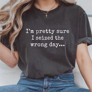 May include: Dark gray t-shirt with the text "I'm pretty sure I seized the wrong day..." in white. The shirt is a crew neck style and is worn with blue jeans. A gold necklace with a cross pendant is visible.