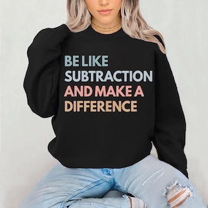 May include: Black sweatshirt with the text "BE LIKE SUBTRACTION AND MAKE A DIFFERENCE" in light blue, pink, and tan. The sweatshirt is a crew neck style.