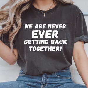 May include: Dark gray t-shirt with the text "WE ARE NEVER EVER GETTING BACK TOGETHER!" in white block letters. The shirt is a crew neck style and appears to be made of a soft, comfortable material. The shirt is paired with blue jeans.