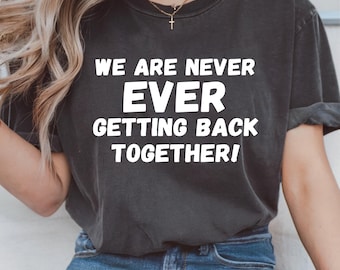 Könnte beinhalten: Dunkelgraues T-Shirt mit dem weißen Text "WE ARE NEVER EVER GETTING BACK TOGETHER!". Das T-Shirt hat einen Rundhalsausschnitt und scheint aus einem weichen, bequemen Material zu sein. Das T-Shirt wird mit einer blauen Jeans getragen.