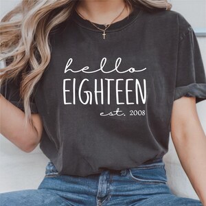 Puede incluir: Camiseta gris oscuro con las palabras "hello EIGHTEEN est. 2008" en blanco. La camiseta tiene cuello redondo y mangas cortas. La modelo lleva vaqueros azules.