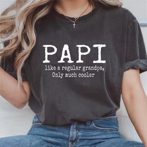 May include: Dark gray t-shirt with the word "PAPI" in large white letters. Below the word "PAPI" is the text "like a regular grandpa, Only much cooler". The shirt is worn with blue jeans.