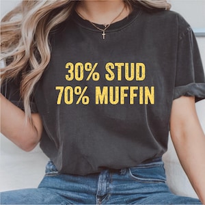 Pode incluir: T-shirt cinza escuro com o texto "30% STUD 70% MUFFIN" em amarelo, com letras desgastadas. A camisa é de gola redonda e parece ser feita de um material macio e confortável. A modelo está usando jeans azuis.