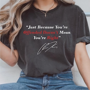 Op de afbeelding: Donkergrijs t-shirt met de quote "Just Because You're Offended Doesn't Mean You're Right" in witte en rode tekst. Het shirt heeft ook een witte handtekening. Het model draagt een blauwe jeans.