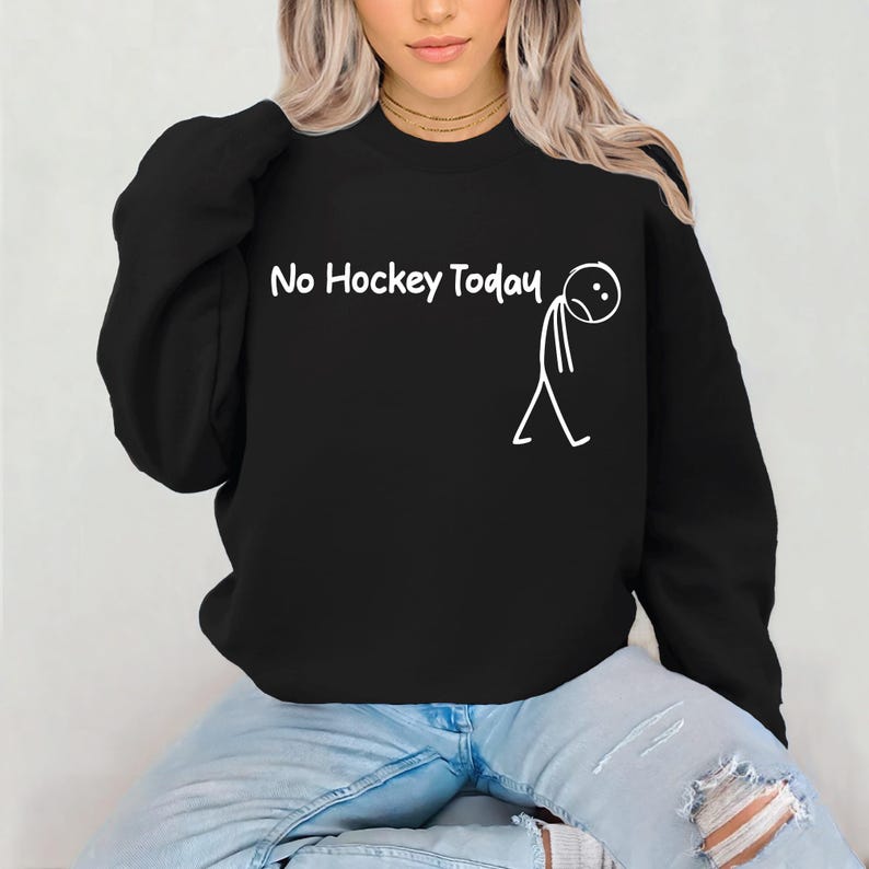 May include: Black sweatshirt with the text "No Hockey Today" in white, accompanied by a simple stick figure with a sad face. The sweatshirt is worn by a person in blue jeans.