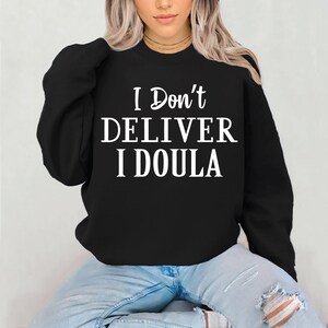 May include: Black sweatshirt with the white text "I Don't DELIVER I DOULA". The sweatshirt is a crew neck style. The model is wearing blue jeans.