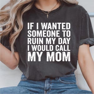 Może przedstawiać: Ciemnoszara koszulka z białym napisem: „JEŚLI CHCIAŁABYM, ŻEBY KTOŚ ZRUJNOWAŁ MI DZIEŃ, ZADZWONIŁABYM DO MAMY”. Koszulka jest noszona przez osobę noszącą niebieskie dżinsy i złoty naszyjnik.