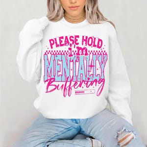 May include: White sweatshirt with the text "PLEASE HOLD I'M MENTALLY BUFFERING" in pink and blue. The sweatshirt has a crew neck and long sleeves. The text is in a bold, playful font.